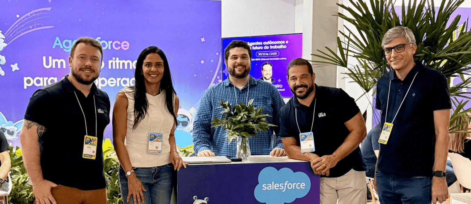 Equipe da ília no stand da Salesforce durante o BB Digital Week 2025. A foto mostra cinco colaboradores da ília, incluindo quatro homens e uma mulher. Eles estão posicionados em frente a um balcão azul com a marca 'Salesforce' e um mascote robótico. Ao fundo, um telão exibe o tema 'Agentes autônomos e futuro do trabalho', refletindo a discussão sobre IA e inovação no evento. A presença da equipe destaca a parceria da ília com a Salesforce e seu engajamento com as tendências em tecnologia e finanças.