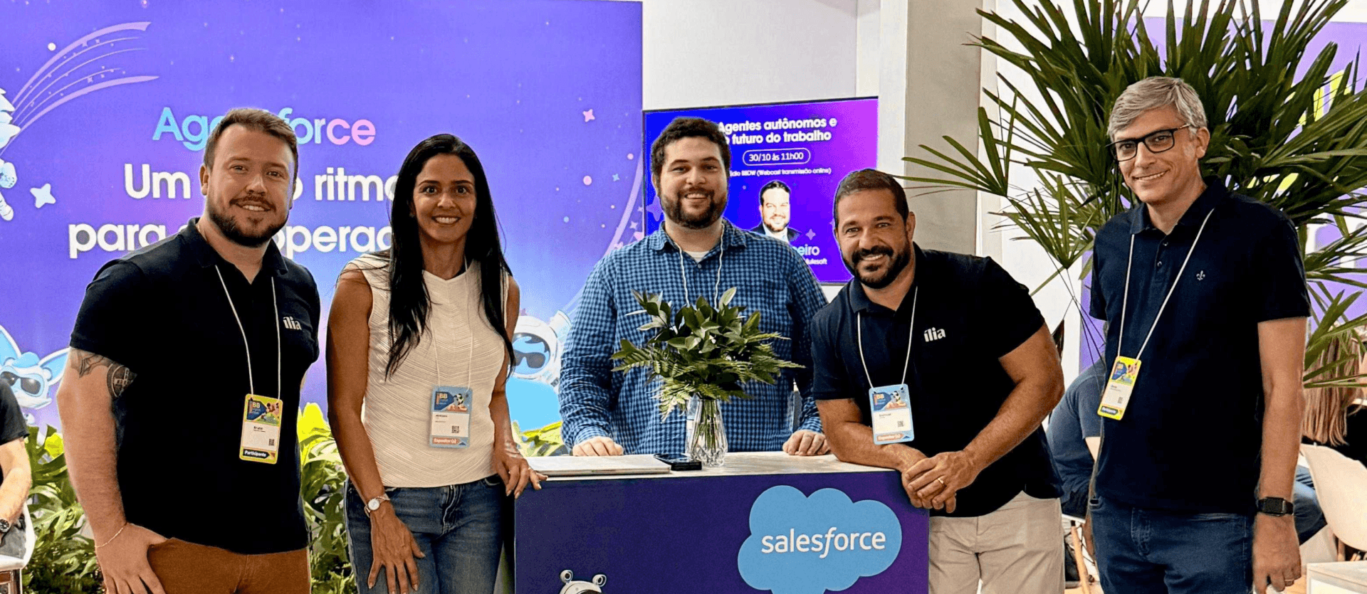 Equipe da ília no stand da Salesforce durante o BB Digital Week 2025. A foto mostra cinco colaboradores da ília, incluindo quatro homens e uma mulher. Eles estão posicionados em frente a um balcão azul com a marca 'Salesforce' e um mascote robótico. Ao fundo, um telão exibe o tema 'Agentes autônomos e futuro do trabalho', refletindo a discussão sobre IA e inovação no evento. A presença da equipe destaca a parceria da ília com a Salesforce e seu engajamento com as tendências em tecnologia e finanças.
