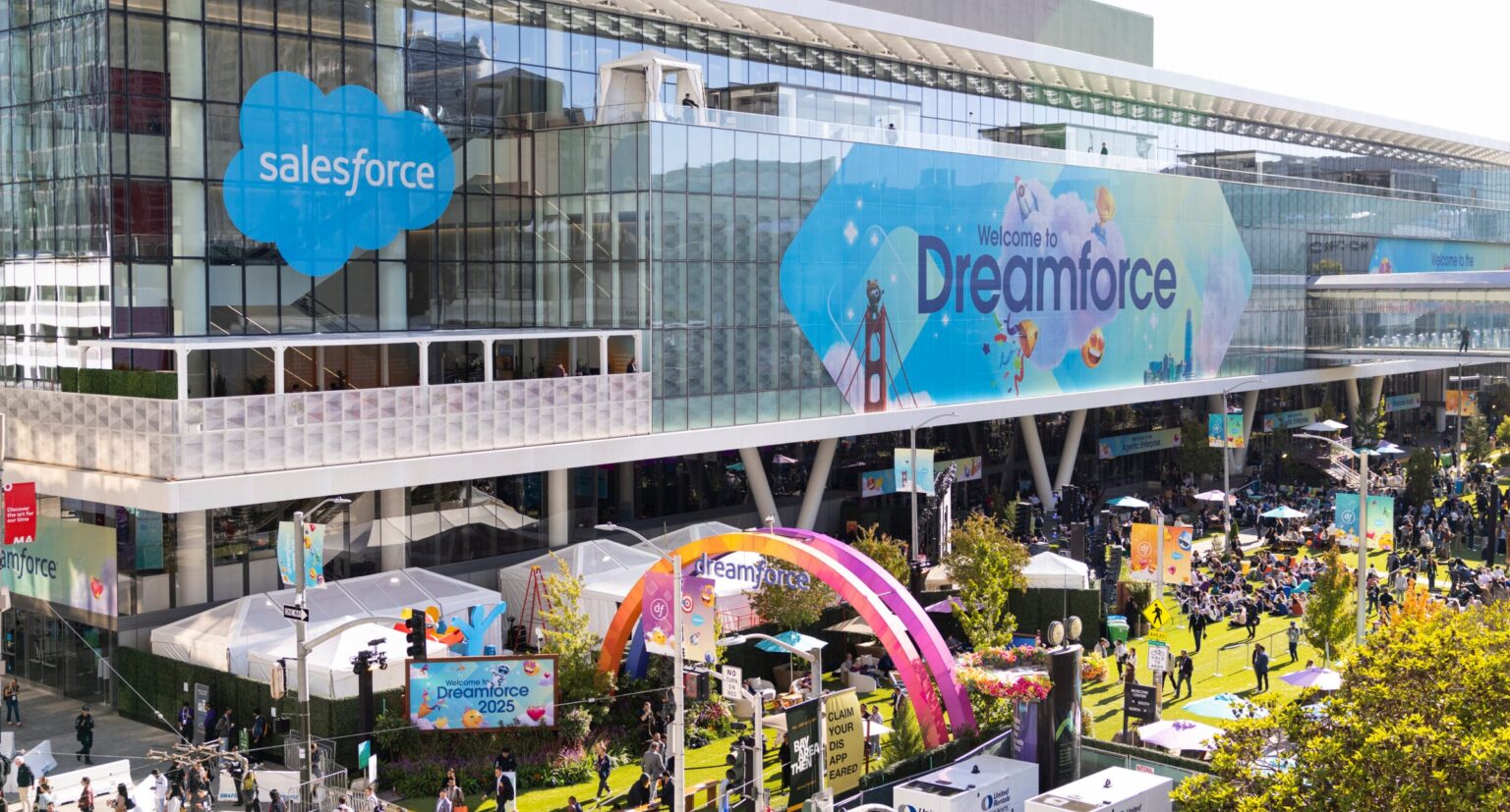 Foto oficial da Salesforce do Dreamforce 2025, realizada no Moscone Center, em São Francisco. A imagem mostra o arco colorido de entrada, o logotipo da empresa e o público participando das atividades no espaço externo do evento.