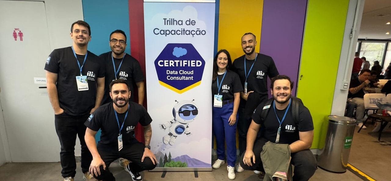 Grupo de seis ílians posando ao lado de um banner com a inscrição 'Trilha de Capacitação Certified Data Cloud Consultant'. Eles vestem camisetas pretas com o logo da ília, posando em ambiente interno colorido e moderno, simbolizando o compromisso da equipe com o desenvolvimento contínuo e a certificação técnica.