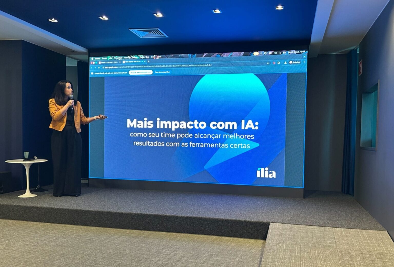 Luciana Oliveira, Head de Design e Experiência na Ília Digital, apresenta palestra sobre IA e impacto nos resultados de equipes, em auditório moderno, com slide: 'Mais impacto com IA: como seu time pode alcançar melhores resultados com as ferramentas certas'.