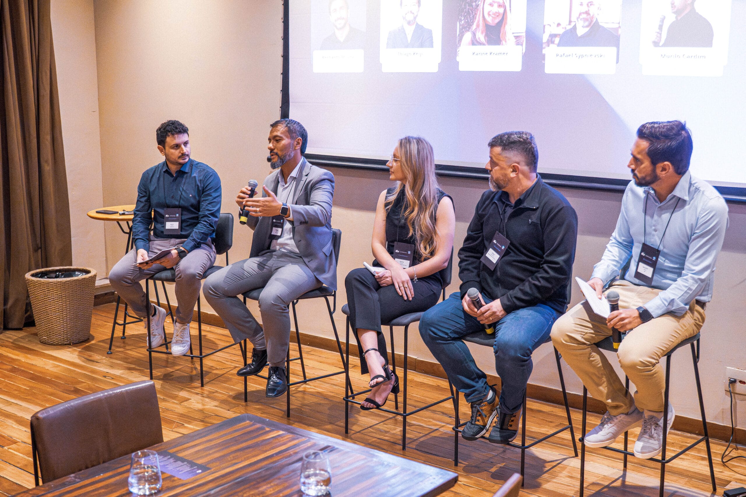Painel de especialistas no evento ília Experience, com cinco profissionais sentados em bancos altos e falando em microfones. Ao fundo, um telão exibe fotos e nomes dos palestrantes.